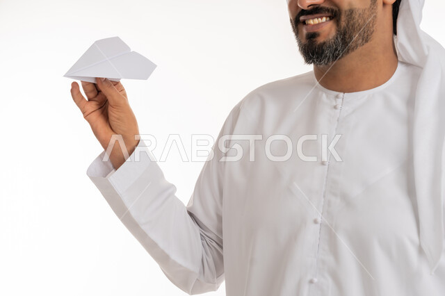 تطوير الأفكار و التطلع للمستقبل, التحقيق والوصول الى الهدف, النظر الى الكاميرا بايماءات الفرح والسرور، بورتريه مقرب لرجل عربي خليجي اماراتي مبتسم يرتدي الكندورة والغترة الاماراتية يحمل طائرة مصنوعة من الورق, خلفية بيضاء