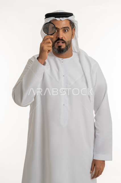 توضيح الصورة والرؤية عن قرب، مفهوم الفحص والاستكشاف، بورتريه لرجل عربي خليجي اماراتي يرتدي الكندورة والغترة الاماراتية ينظر من العدسة المكبرة بإيماءات المفاجئة والصدمة،  خلفية بيضاء