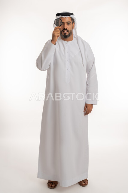 مفهوم الفحص والاستكشاف، توضيح الصورة والرؤية عن قرب، بورتريه لرجل عربي خليجي اماراتي يرتدي الكندورة والغترة الاماراتية ينظر من العدسة المكبرة بإيماءات المفاجئة والصدمة، الجسم الكامل بالطول، خلفية بيضاء