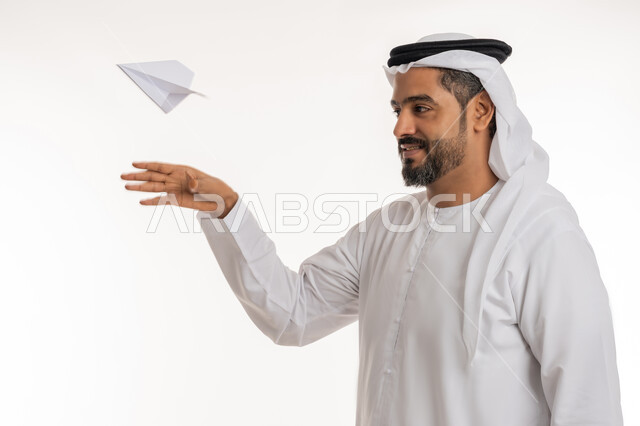 التحقيق والوصول الى الهدف, تطوير الأفكار و التطلع للمستقبل, بورتريه مقرب لرجل عربي خليجي اماراتي مبتسم يرتدي الكندورة والغترة الاماراتية يقوم برمي طائرة أوريغامي بأجنحة مصنوعة من الورق, خلفية بيضاء