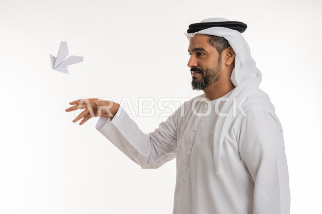 التحقيق والوصول الى الهدف, تطوير الأفكار و التطلع للمستقبل, بورتريه مقرب لرجل عربي خليجي اماراتي مبتسم يرتدي الكندورة والغترة الاماراتية يقوم برمي طائرة أوريغامي بأجنحة مصنوعة من الورق, خلفية بيضاء