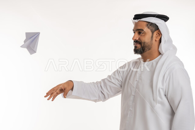 Investigation and reaching the goal, developing ideas and looking forward to the future, close-up portrait of a smiling Emirati Gulf Arab man wearing a kandura and an Emirati ghutra throwing an origami plane with wings made of paper, white background