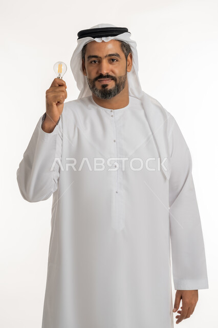 أفكار ابداعية تطويرية، تحقيق الانجازات والابتكارات، بورتريه لرجل عربي خليجي اماراتي مبتسم يرتدي الكندورة والغترة يحمل بيده المصباح الكهربائي الضوئي، النظر الى الكاميرا بتعابيرالفخر والسرور، براعة الاختراع والمهارة والقدرات الذهنية، خلفية بيضاء