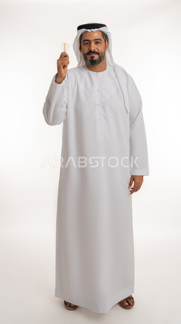 مفهوم الفكرة مع الإبتكار والإلهام، أفكار ابداعية تطويرية، تحقيق الانجازات والابتكارات، بورتريه لرجل عربي خليجي اماراتي مبتسم يرتدي الكندورة والغترة يحمل بيده المصباح الكهربائي الضوئي، النظر الى الكاميرا بتعابيرالفخر والسرور والثقة بالنفس، الجسم الكامل بالطول، خلفية بيضاء