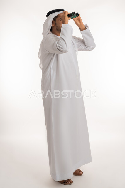 Adventure and love of exploration, future planning for goals and ambitions, seeing distant things, portrait of a smiling Emirati Gulf Arab man wearing a kandura and a ghutra holding binoculars in his hand looking at something with gestures of joy and pleasure, full body image, white background