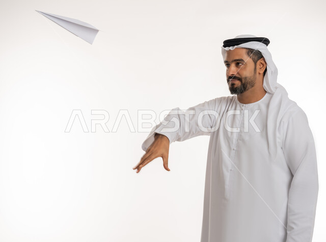التحقيق والوصول الى الهدف, تطوير الأفكار و التطلع للمستقبل, بورتريه مقرب لرجل عربي خليجي اماراتي مبتسم يرتدي الكندورة والغترة الاماراتية يقوم برمي طائرة أوريغامي بأجنحة مصنوعة من الورق, خلفية بيضاء