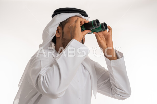 رؤية الاشياء البعيدة، التخطيط المستقبلي للأهداف والطموحات، المغامرة و حب الاستكشاف، بورتريه مقرب لرجل عربي خليجي اماراتي مبتسم يرتدي الكندورة والغترة يحمل بيده المنظار ثنائي العينين ينظر الى شيء ما بإيماءات الفرح و السرور، خلفية بيضاء