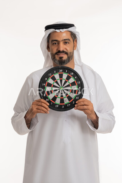 النظر الى الكاميرا بتعابير السرور, مهارات رياضية و تسليه وترفيه، بورتريه لرجل عربي خليجي اماراتي مبتسم يرتدي الكندورة والغترة الاماراتية يحمل لعبة السهام المغناطيسية، مفهوم التركيز وتحديد الأهداف, خلفية بيضاء