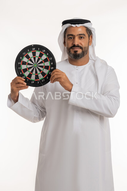 النظر الى الكاميرا بتعابير السرور, مهارات رياضية و تسليه وترفيه، بورتريه لرجل عربي خليجي اماراتي مبتسم يرتدي الكندورة والغترة الاماراتية يحمل لعبة السهام المغناطيسية، مفهوم التركيز وتحديد الأهداف, خلفية بيضاء