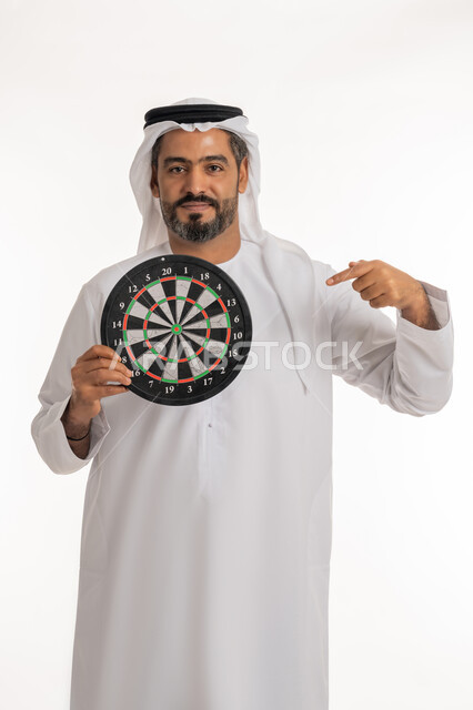 تسليه وترفيه و مهارات رياضية، مفهوم التركيز وتحديد الأهداف, النظر الى الكاميرا بتعابير السرور, بورتريه لرجل عربي خليجي اماراتي مبتسم يرتدي الكندورة والغترة الاماراتية يشير باصبع السبابة الى لعبة السهام المغناطيسية، خلفية بيضاء