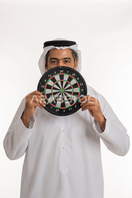 مفهوم التركيز وتحديد الأهداف, النظر الى الكاميرا بتعابير السرور, مهارات رياضية و تسليه وترفيه، بورتريه لرجل عربي خليجي اماراتي مبتسم يرتدي الكندورة والغترة الاماراتية يحمل لعبة السهام المغناطيسية، خلفية بيضاء