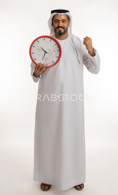 انجاز المهامات في الوقت المناسب، النظر الى الكاميرا بتعابير الفرح والسرور، بورتريه لرجل عربي خليجي اماراتي مبتسم يرتدي الكندورة والغترة الاماراتية يحمل بيده ساعة رقمية كبيرة يرفع قبضة يده للاعلى، التذكيرات والتنبيهات الثابته، مفهوم العد التنازلي، خلفية بيضاء