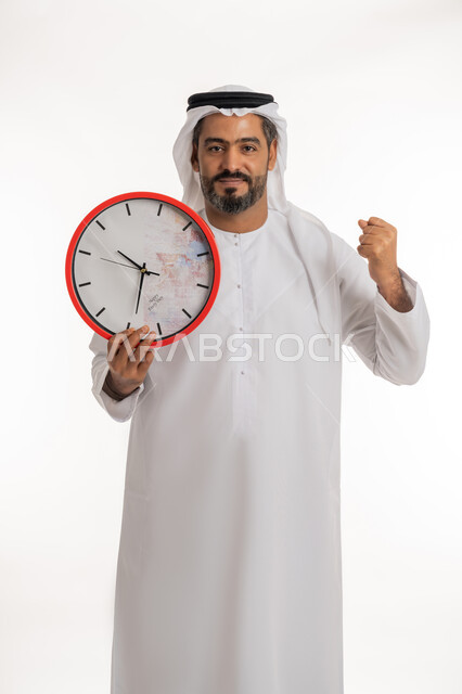 النظر الى الكاميرا بتعابير الفرح والسرور، انجاز المهامات في الوقت، بورتريه لرجل عربي خليجي اماراتي مبتسم يرتدي الكندورة والغترة الاماراتية يحمل بيده ساعة رقمية كبيرة يرفع قبضة يده للاعلى ، التذكيرات والتنبيهات الثابته، مفهوم العد التنازلي، خلفية بيضاء