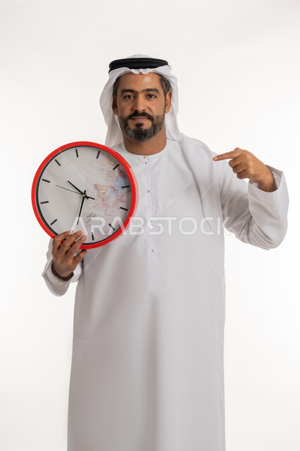 انجاز المهامات في الوقت، بورتريه لرجل عربي خليجي اماراتي مبتسم يرتدي الكندورة والغترة الاماراتية يحمل بيده ساعة رقمية كبيرة يشير بإصبع السبابة الى التوقيت،  النظر الى الكاميرا بتعابير الفرح والسرور، الدقة في المواعيد ، التذكيرات والتنبيهات الثابته، مفهوم العد التنازلي، خلفية بيضاء
