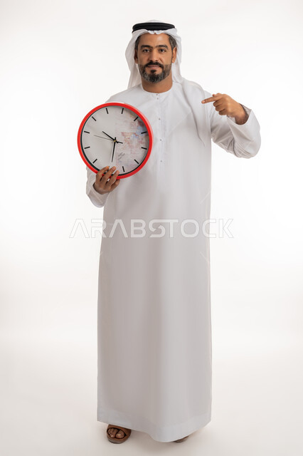مفهوم العد التنازلي، انجاز المهامات في الوقت، بورتريه لرجل عربي خليجي اماراتي مبتسم يرتدي الكندورة والغترة الاماراتية يحمل بيده ساعة رقمية كبيرة يشير بإصبع السبابة الى التوقيت،  النظر الى الكاميرا بتعابير الفرح والسرو، التذكيرات والتنبيهات الثابته، الجسم الكامل بالطول، خلفية بيضاء