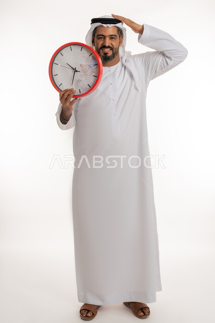 النظر الى الكاميرا بتعابير الفرح والسرور، انجاز المهامات في الوقت، بورتريه لرجل عربي خليجي اماراتي مبتسم يرتدي الكندورة والغترة الاماراتية يحمل بيده ساعة رقمية كبيرة يضع يده على رأسه، التذكيرات والتنبيهات الثابته، مفهوم العد التنازلي، الجسم الكامل بالطول، خلفية بيضاء