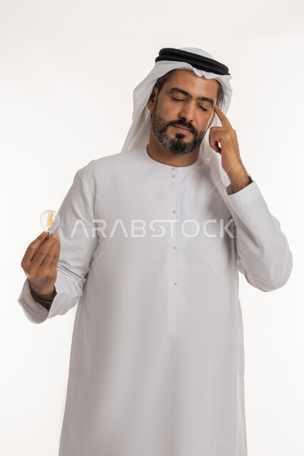 Closing eyes and placing index finger on head with expressions of thinking, contemplation and concentration, creative development ideas, portrait of an Arab Gulf Emirati man wearing a kandura and a ghutra holding an electric light bulb in his hand, the concept of an idea with innovation and inspiration, achieving accomplishments and innovations, white background