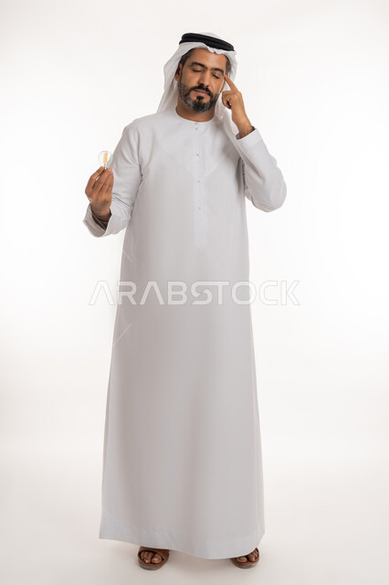 اغماض العينين ووضع اصبع السبابة على الرأس بتعابير التفكير والتمعن والتركيز، أفكار ابداعية تطويرية، بورتريه لرجل عربي خليجي اماراتي يرتدي الكندورة والغترة يحمل بيده المصباح الكهربائي الضوئي، مفهوم الفكرة مع الإبتكار والإلهام، الجسم الكامل بالطول، خلفية بيضاء