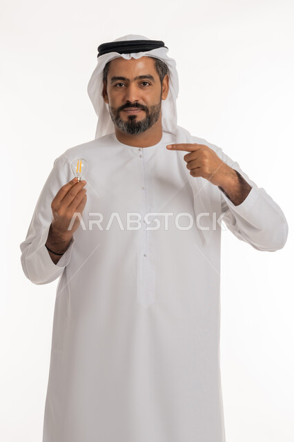 النظر الى الكاميرا والاشارة بإصبع السبابة الى الاختراع، أفكار ابداعية تطويرية، بورتريه لرجل عربي خليجي اماراتي يرتدي الكندورة والغترة يحمل بيده المصباح الكهربائي الضوئي بإيماءات الفخر والسرور، مفهوم الفكرة مع الإبتكار والإلهام، تحقيق الانجازات والابتكارات، خلفية بيضاء