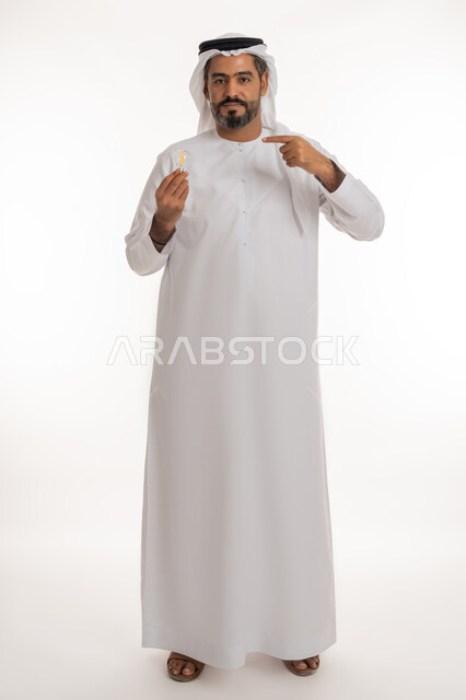 تحقيق الانجازات والابتكارات، النظر الى الكاميرا والاشارة بإصبع السبابة الى الاختراع، أفكار ابداعية تطويرية، بورتريه لرجل عربي خليجي اماراتي يرتدي الكندورة والغترة يحمل بيده المصباح الكهربائي الضوئي بإيماءات الفخر والسرور، مفهوم الفكرة مع الإبتكار والإلهام، الجسم الكامل بالطول خلفية بيضاء