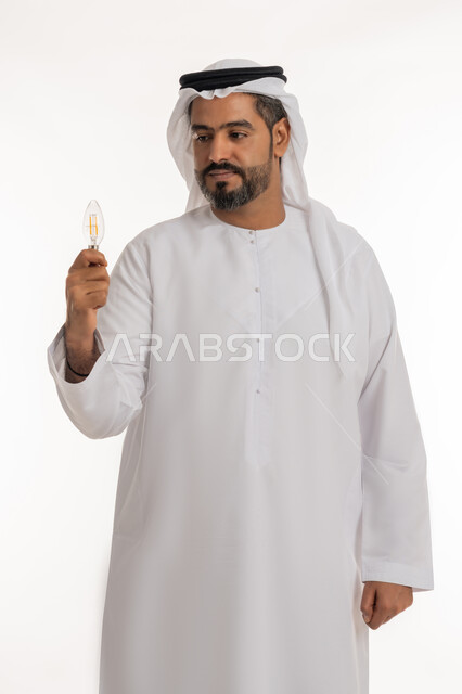 براعة المهارات والقدرات الذهنية، تحقيق الانجازات والابتكارات، بورتريه لرجل عربي خليجي اماراتي يرتدي الكندورة والغترة يحمل بيده المصباح الكهربائي الضوئي، النظر الى الاخترع بتعابيرالتفكير والفخر، مفهوم الفكرة مع الإبتكار والإلهام، أفكار ابداعية تطويرية، خلفية بيضاء