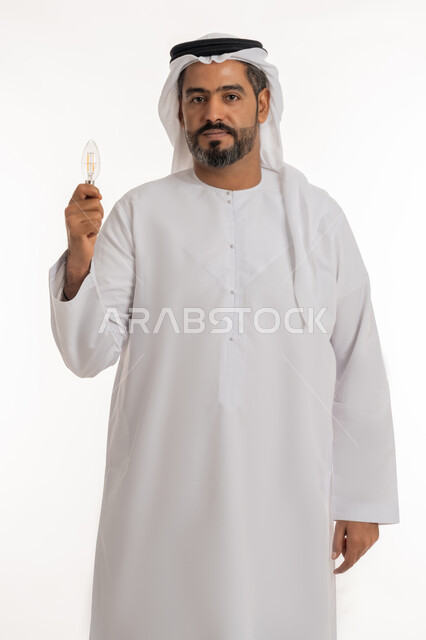 أفكار ابداعية تطويرية، تحقيق الانجازات والابتكارات، بورتريه لرجل عربي خليجي اماراتي مبتسم يرتدي الكندورة والغترة يحمل بيده المصباح الكهربائي الضوئي، النظر الى الكاميرا بتعابيرالفخر والسرور والثقة بالنفس، مفهوم الفكرة مع الإبتكار والإلهام، براعة الاختراع والمهارة والقدرات الذهنية، خلفية بيضاء