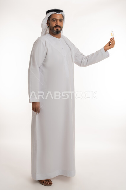 Concept of idea with innovation and inspiration, creative development ideas, achieving accomplishments and innovations, portrait of a smiling Emirati Gulf Arab man wearing a kandura and a ghutra holding a light bulb in his hand, looking at the camera with expressions of pride, joy and self-confidence, full body length, white background