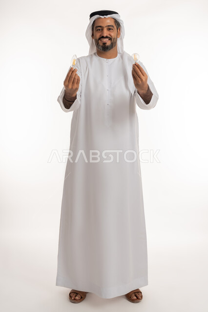 مفهوم الفكرة مع الإبتكار والإلهام، أفكار ابداعية تطويرية، تحقيق الانجازات والابتكارات، بورتريه لرجل عربي خليجي اماراتي مبتسم يرتدي الكندورة والغترة يحمل بيده المصباح الكهربائي الضوئي، النظر الى الكاميرا بتعابيرالفخر والسرور والثقة بالنفس، الجسم الكامل بالطول، خلفية بيضاء