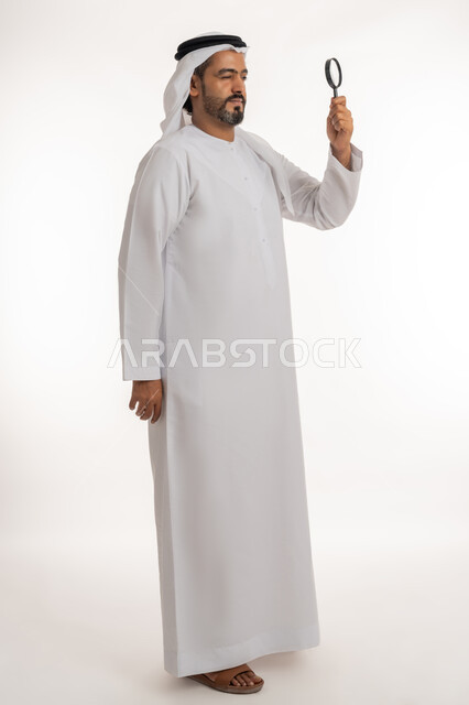 مفهوم الفحص والاستكشاف، توضيح الصورة والرؤية عن قرب، بورتريه من الجانب لرجل عربي خليجي اماراتي يرتدي الكندورة والغترة الاماراتية ينظر من العدسة مكبرة بإيماءات التركيز والتمعن، الجسم الكامل بالطول، خلفية بيضاء
