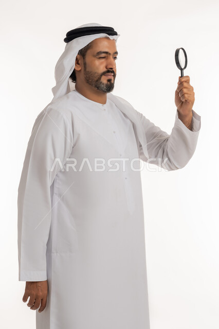 توضيح الصورة والرؤية عن قرب، بورتريه من الجانب لرجل عربي خليجي اماراتي يرتدي الكندورة والغترة الاماراتية يحمل بيده عدسة مكبرة بإيماءات التركيز والتمعن، مفهوم الفحص والاستكشاف، خلفية بيضاء