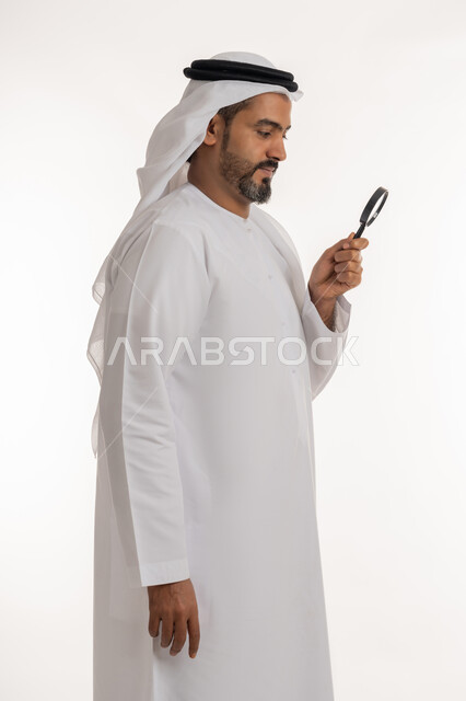 مفهوم الفحص والاستكشاف، توضيح الصورة والرؤية عن قرب، بورتريه من الجانب لرجل عربي خليجي اماراتي يرتدي الكندورة والغترة الاماراتية ينظر من العدسة مكبرة بإيماءات التركيز والتمعن، الجسم الكامل بالطول، خلفية بيضاء