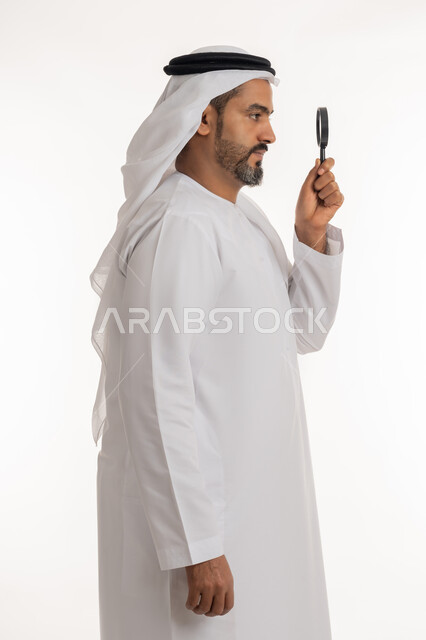توضيح الصورة والرؤية عن قرب، بورتريه من الجانب لرجل عربي خليجي اماراتي يرتدي الكندورة والغترة الاماراتية يحمل بيده عدسة مكبرة بإيماءات التركيز والتمعن، مفهوم الفحص والاستكشاف، خلفية بيضاء