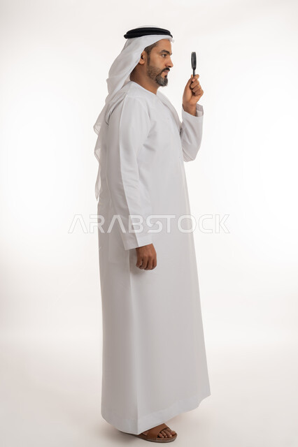 مفهوم الفحص والاستكشاف، توضيح الصورة والرؤية عن قرب، بورتريه من الجانب لرجل عربي خليجي اماراتي يرتدي الكندورة والغترة الاماراتية ينظر من العدسة مكبرة بإيماءات التركيز والتمعن، الجسم الكامل بالطول، خلفية بيضاء