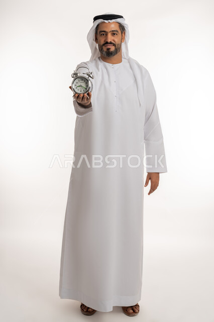 إنجاز المهامات في الوقت المحدد، مفهوم تنظيم وإدارة الوقت، بورتريه لرجل عربي خليجي اماراتي يرتدي الكندورة والغترة الاماراتية يحمل ساعة منبه صغيرة بتعابير الفخر والسرور ، الدقة في مواعيد العمل، التذكيرات والتنبيهات الثابته، الجسم الكامل بالطول، خلفية بيضاء