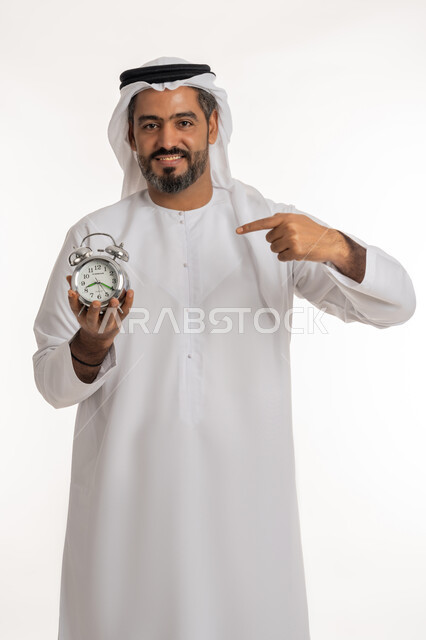 الدقة في مواعيد العمل، مفهوم تنظيم وإدارة الوقت، بورتريه لرجل عربي خليجي اماراتي مبتسم يرتدي الكندورة والغترة الاماراتية يشير بإصبع السبابة الى ساعة المنبه بإيماءات السرور والفرح، التذكيرات والتنبيهات الثابته، إنجاز المهامات في الوقت المحدد، خلفية بيضاء