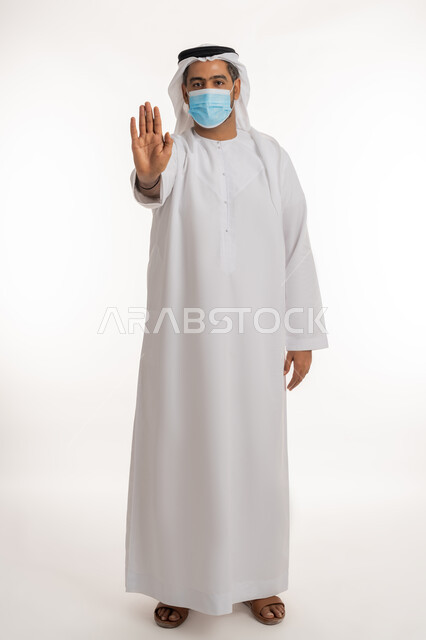 Prohibition of doing something, following the Ministry of Health's precautionary measures, the importance of receiving the Corona virus vaccination, spreading health awareness to reduce the spread of diseases, portrait of an Arab Gulf Emirati man wearing an Emirati kandura and ghutra raising his palm up with gestures of rejection, dissatisfaction and acceptance, full body lengthwise, white background