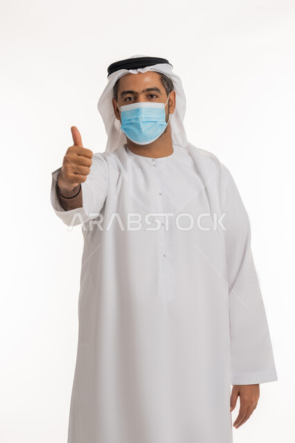 اتباع إجراءات وزارة الصحة الاحترازية، أهمية تلقي التطعيم ضد فايروس كورونا، نشر الوعي الصحي للحد من انتشار الأمراض، بورتريه لرجل عربي خليجي إماراتي يرتدي الكندورة وكمامة الوقاية يرفع اصبعه الابهام لاعلى بإيماءات القبول والإعجاب، خلفية بيضاء