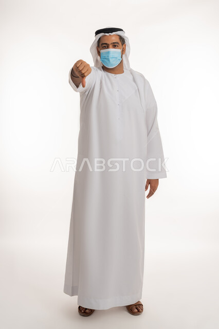 نشر الوعي الصحي للحد من انتشار الأمراض، أهمية تلقي التطعيم ضد فايروس كورونا، بورتريه لرجل عربي خليجي إماراتي يرتدي الكندورة وكمامة الوقاية يرفع اصبعه للأسفل بإيماءات الرفض وعدم الإعجاب، اتباع إجراءات وزارة الصحة الاحترازية, الجسم الكامل بالطول، خلفية بيضاء