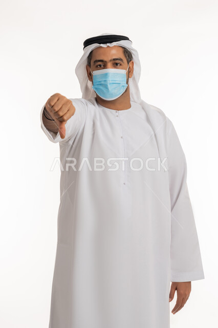 نشر الوعي الصحي للحد من انتشار الأمراض، أهمية تلقي التطعيم ضد فايروس كورونا، بورتريه لرجل عربي خليجي إماراتي يرتدي الكندورة وكمامة الوقاية يرفع اصبعه للأسفل بإيماءات الرفض وعدم الإعجاب، اتباع إجراءات وزارة الصحة الاحترازية, خلفية بيضاء