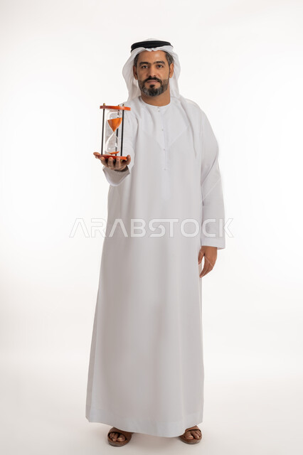 مفهوم إدارة الأعمال وتنظيم الجدول الزمني, إدراك قيمة اللحظات الثمينة, بورتريه لرجل عربي خليجي اماراتي يرتدي الكندورة والغترة يمسك بيده الساعة الرملية الزجاجية (البنكام), النظر إلى الكاميرا بإيماءات السرور, مراقبة توقيت الرمل, صورة لكامل الجسم, خلفية بيضاء