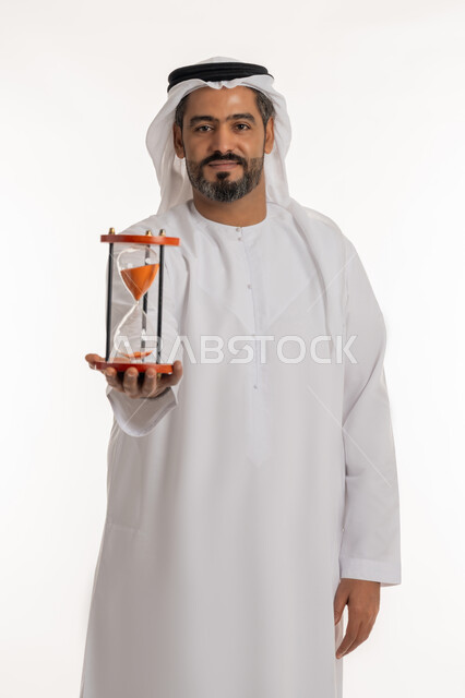 Standing and looking at the camera with gestures of pleasure, doing sand time monitoring, business management and schedule organization concept, realizing the value of precious moments, portrait of a smiling Emirati Gulf Arab man wearing a kandura and ghutra holding a glass sand hourglass (bankam) in his hand, white background