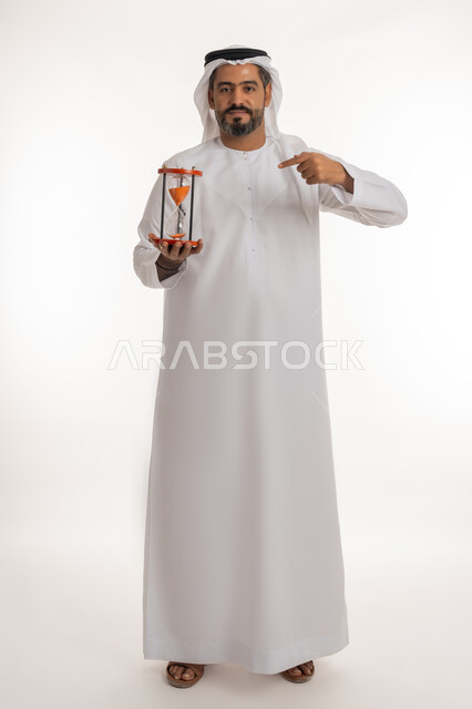 إدراك قيمة اللحظات الثمينة, بورتريه لرجل عربي خليجي اماراتي يرتدي الكندورة والغترة ينظر إلى الكاميرا, الاشارة بإصبع السبابة إلى الساعة الرملية الزجاجية (البنكام), مراقبة توقيت الرمل, مفهوم إدارة الأعمال وتنظيم الجدول الزمني, صورة الجسم بالكامل, خلفية بيضاء