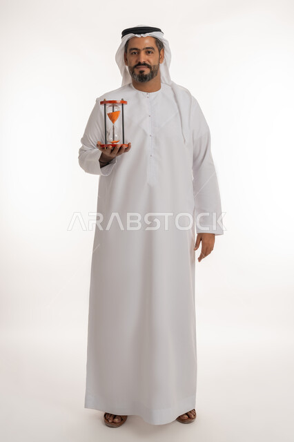 النظر إلى الكاميرا بإيماءات السرور, مراقبة توقيت الرمل, مفهوم إدارة الأعمال وتنظيم الجدول الزمني, إدراك قيمة اللحظات الثمينة, بورتريه لرجل عربي خليجي اماراتي يرتدي الكندورة والغترة يمسك بيده الساعة الرملية الزجاجية (البنكام), صورة لكامل الجسم, خلفية بيضاء