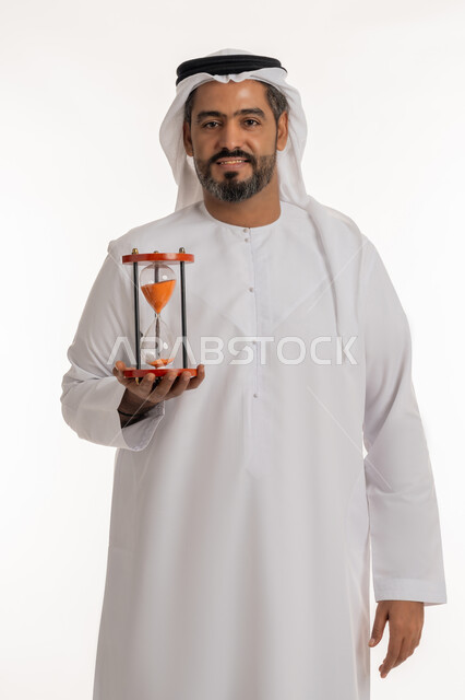 القيام بمراقبة توقيت الرمل, مفهوم إدارة الأعمال وتنظيم الجدول الزمني, إدراك قيمة اللحظات الثمينة, بورتريه لرجل عربي خليجي اماراتي مبتسم يرتدي الكندورة والغترة  يمسك بيده الساعة الرملية الزجاجية (البنكام), النظر إلى الكاميرا بإيماءات السرور, خلفية بيضاء