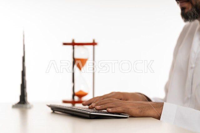 ادراك قيمة الوقت وانجاز المهام بدقة وكفاءة في الزمن المحدد, بورتريه مقرب لرجل عربي خليجي اماراتي يرتدي الكندورة يقوم بالكتابة على لوحة المفاتيح, الدقة في المواعيد، مفهوم ادارة الوقت والجدول الزمني , مجسم ثلاثي الابعاد لبرج خلفية الشهير مع الساعة الرملية، خلفية بيضاء