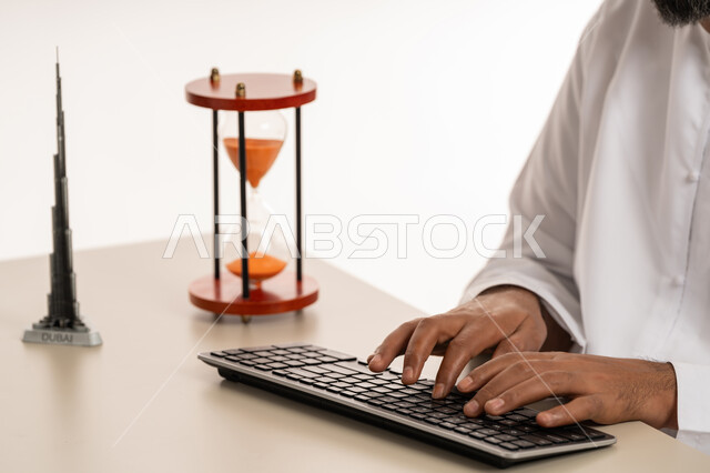 ادراك قيمة الوقت وانجاز المهام بدقة وكفاءة في الزمن المحدد, بورتريه مقرب لرجل عربي خليجي اماراتي يرتدي الكندورة يقوم بالكتابة على لوحة المفاتيح, الدقة في المواعيد، مفهوم ادارة الوقت والجدول الزمني , مجسم ثلاثي الابعاد لبرج خلفية الشهير مع الساعة الرملية، خلفية بيضاء