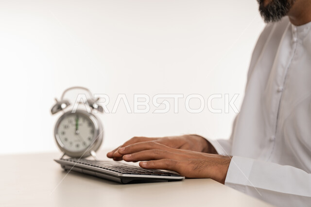 الدقة في المواعيد, مفهوم التذكير والتنبيه, ادراك قيمة الوقت وانجاز المهام في الزمن المحدد, بورتريه مقرب لرجل عربي خليجي اماراتي يرتدي الكندورة يقوم بالكتابة على لوحة المفاتيح, ساعة رقمية صغيرة, خلفية بيضاء