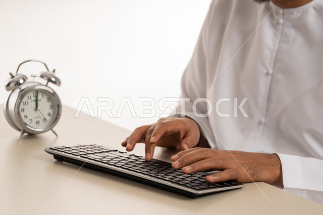 مفهوم التذكير والتنبيه, ادراك قيمة الوقت وانجاز المهام في الزمن المحدد, بورتريه مقرب لرجل عربي خليجي اماراتي يرتدي الكندورة يقوم بالكتابة على لوحة المفاتيح, ساعة رقمية صغيرة,  الدقة في المواعيد, خلفية بيضاء