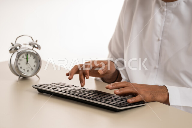The concept of reminder and alert, realizing the value of time and completing tasks on time, close-up portrait of an Arab Gulf Emirati man wearing a kandura typing on a keyboard, small digital watch, punctuality, white background