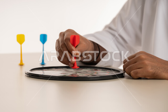 التسلية والترفيه والتحدي في رمي السهام, مفهوم التصويب والتركيز، الدقة في تحديد الهدف، هوايات ومهارات رياضية، بورتريه مقرب ليد رجل عربي خليجي اماراتي يرتدي الكندورة البيضاء يمسك سهم مغناطيسي أحمر اللون, خلفية بيضاء